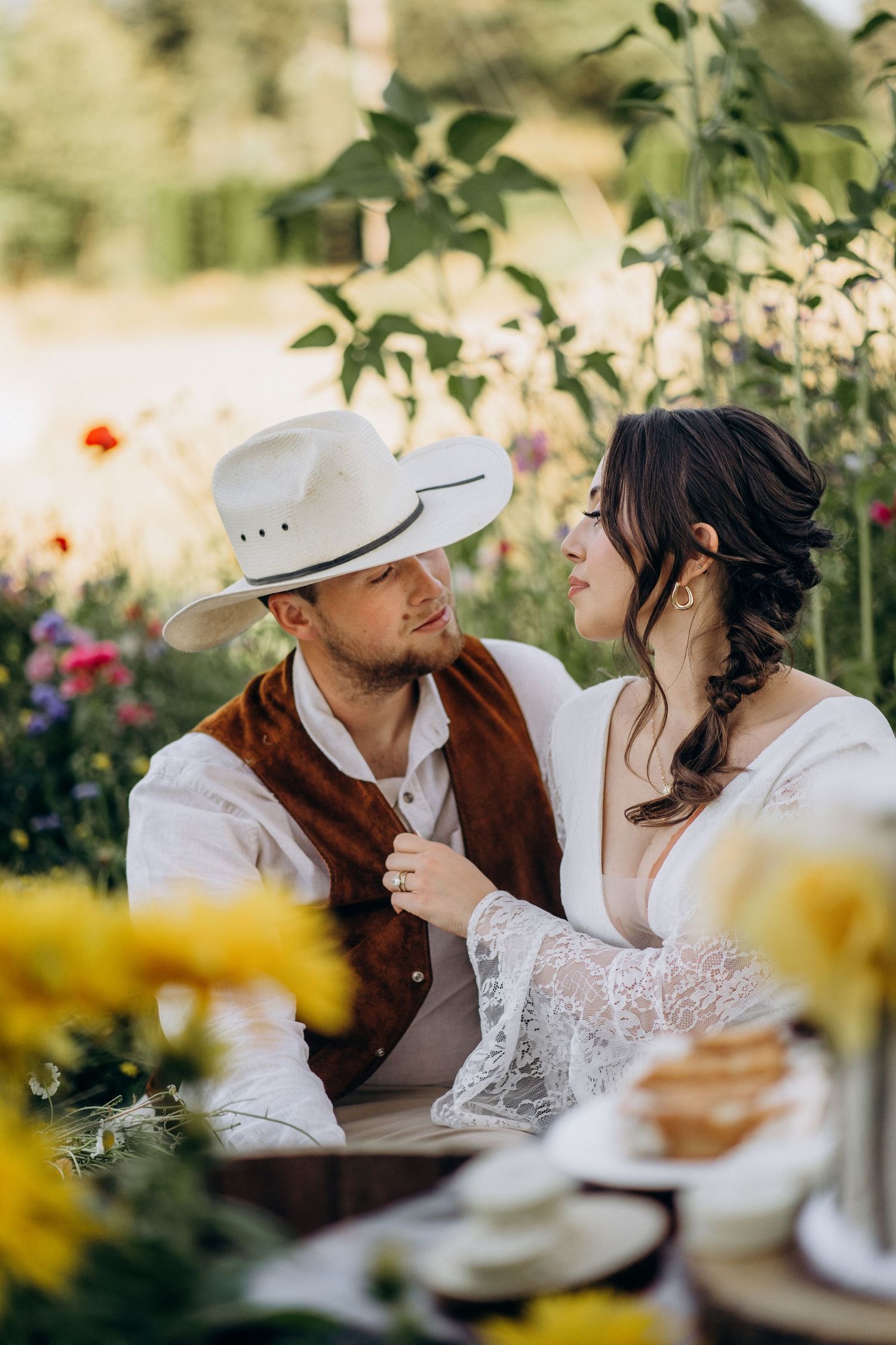 Bride and groom share an intimate moment at a floral garden picnic, groom wearing a white cowboy hat and brown vest.