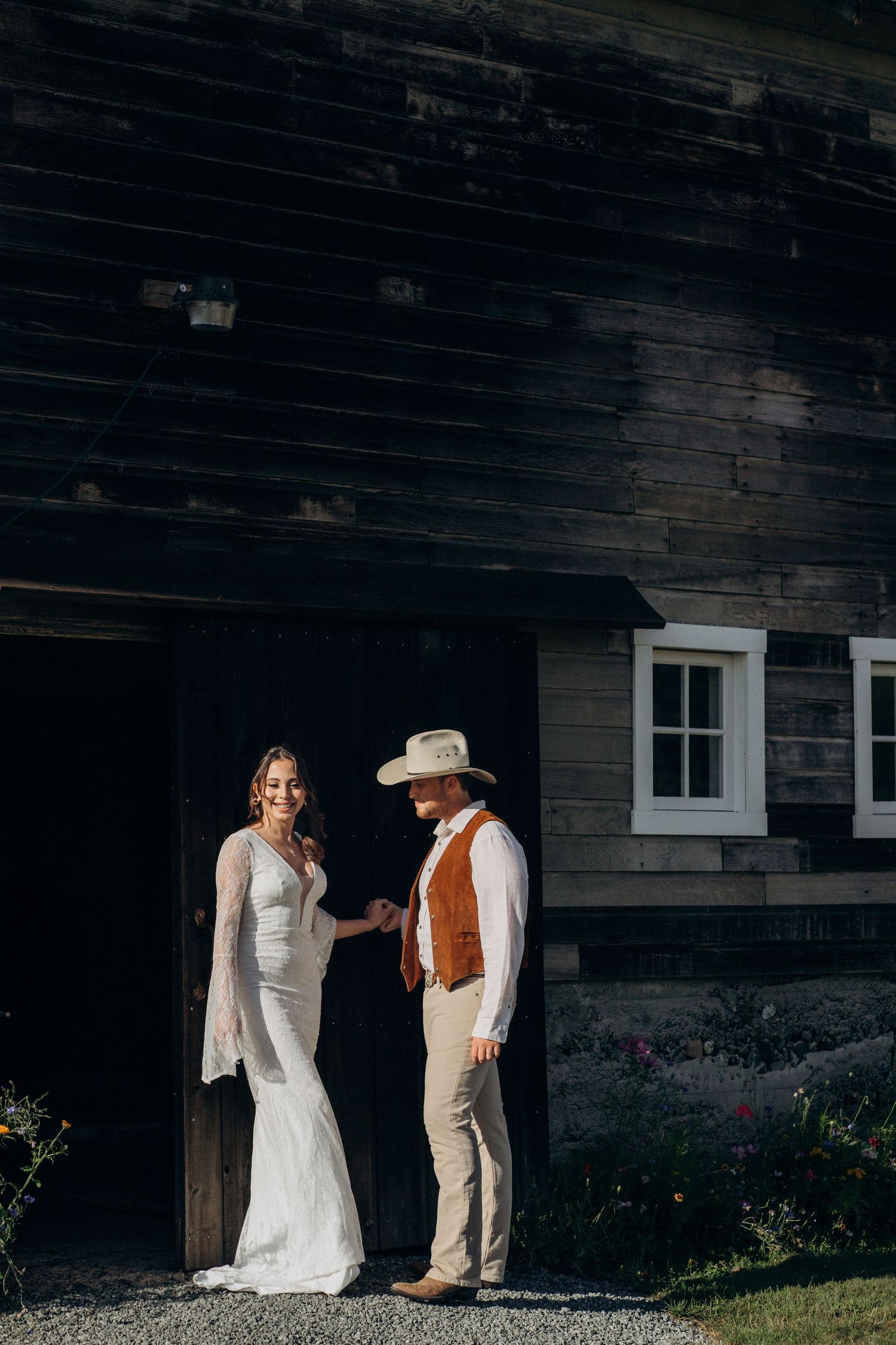 Bride and groom holding hands in golden sunlight outside dark rustic wooden barn.