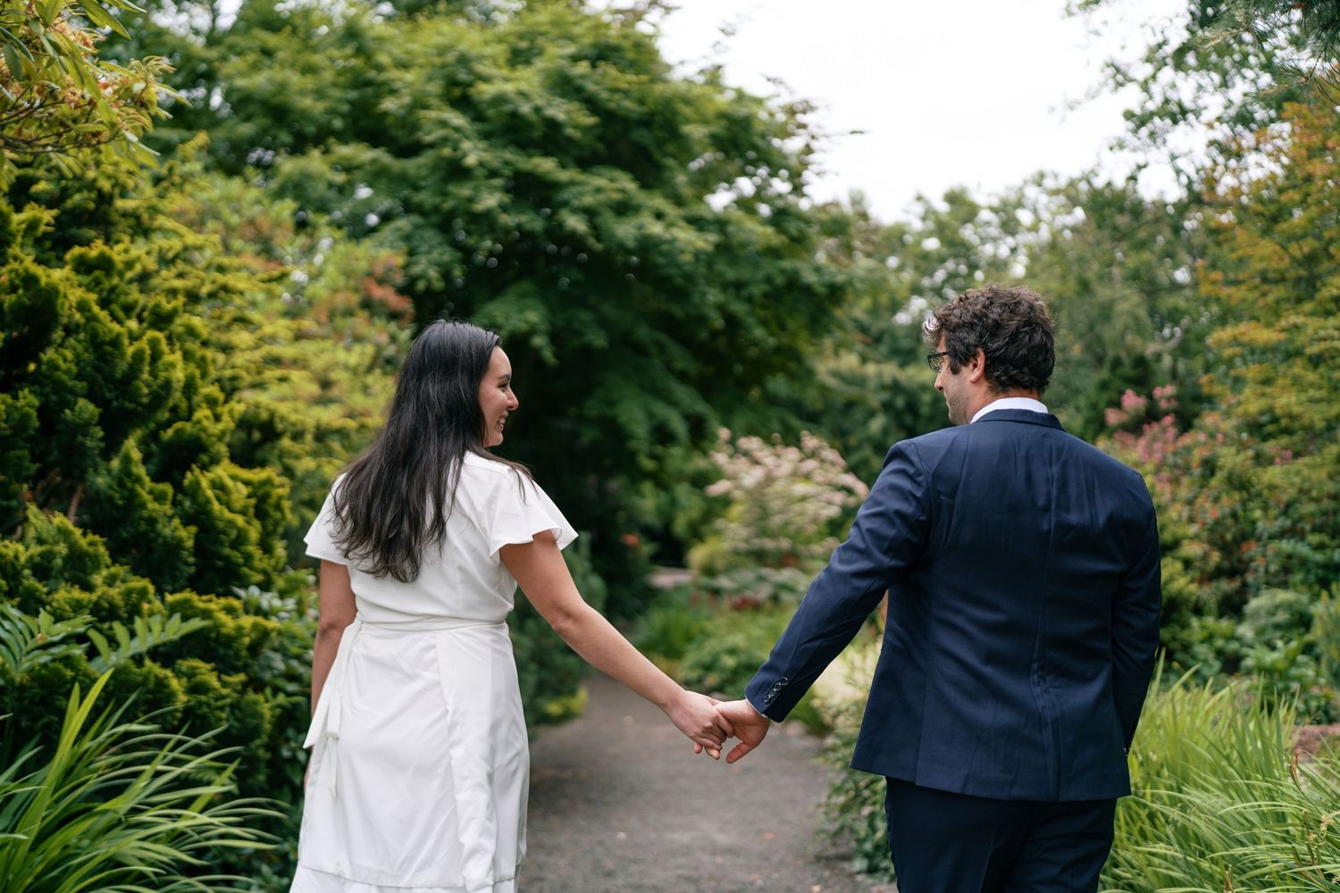 Bride and groom walking hand in hand along a lush green garden path at their wedding.