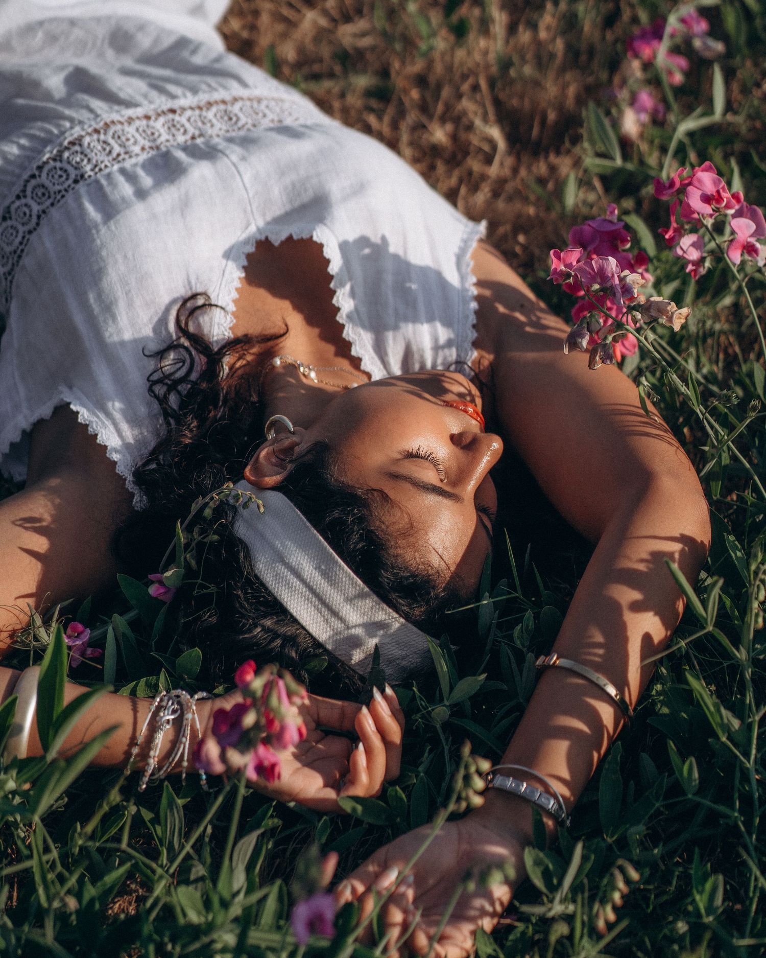 Woman in white dress lying in flower field with eyes closed, surrounded by pink blooms and green grass.