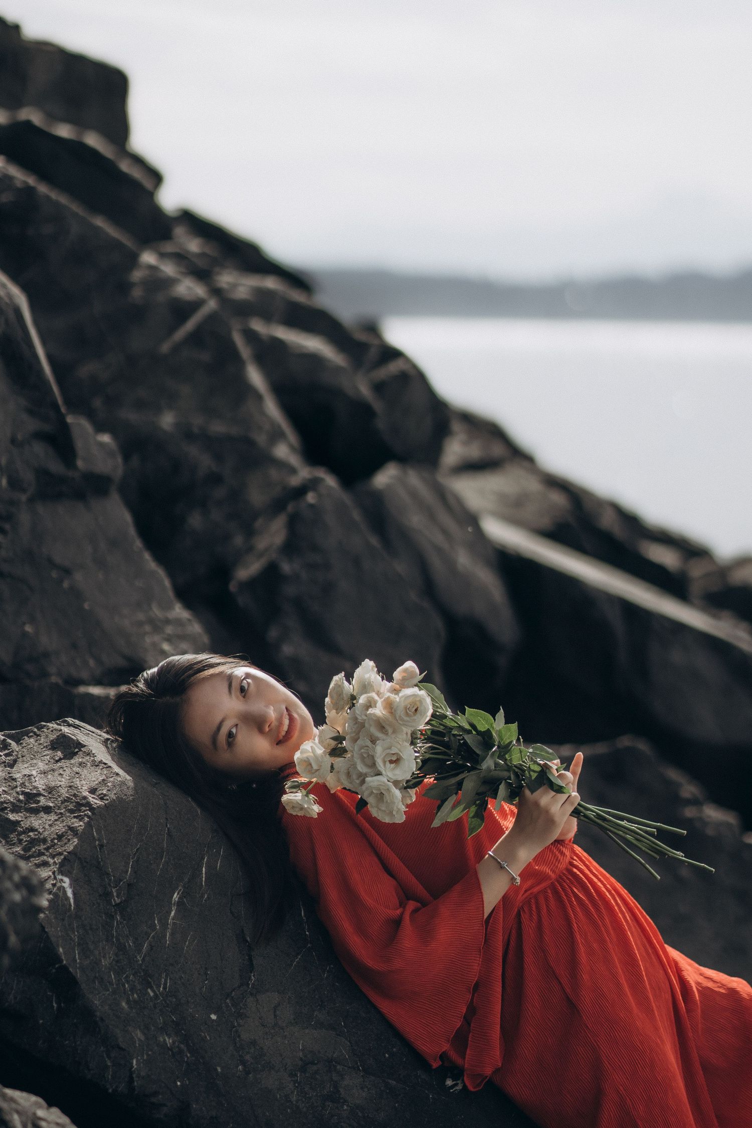 Smiling woman in red dress lying on dark rocks holding white flower bouquet.