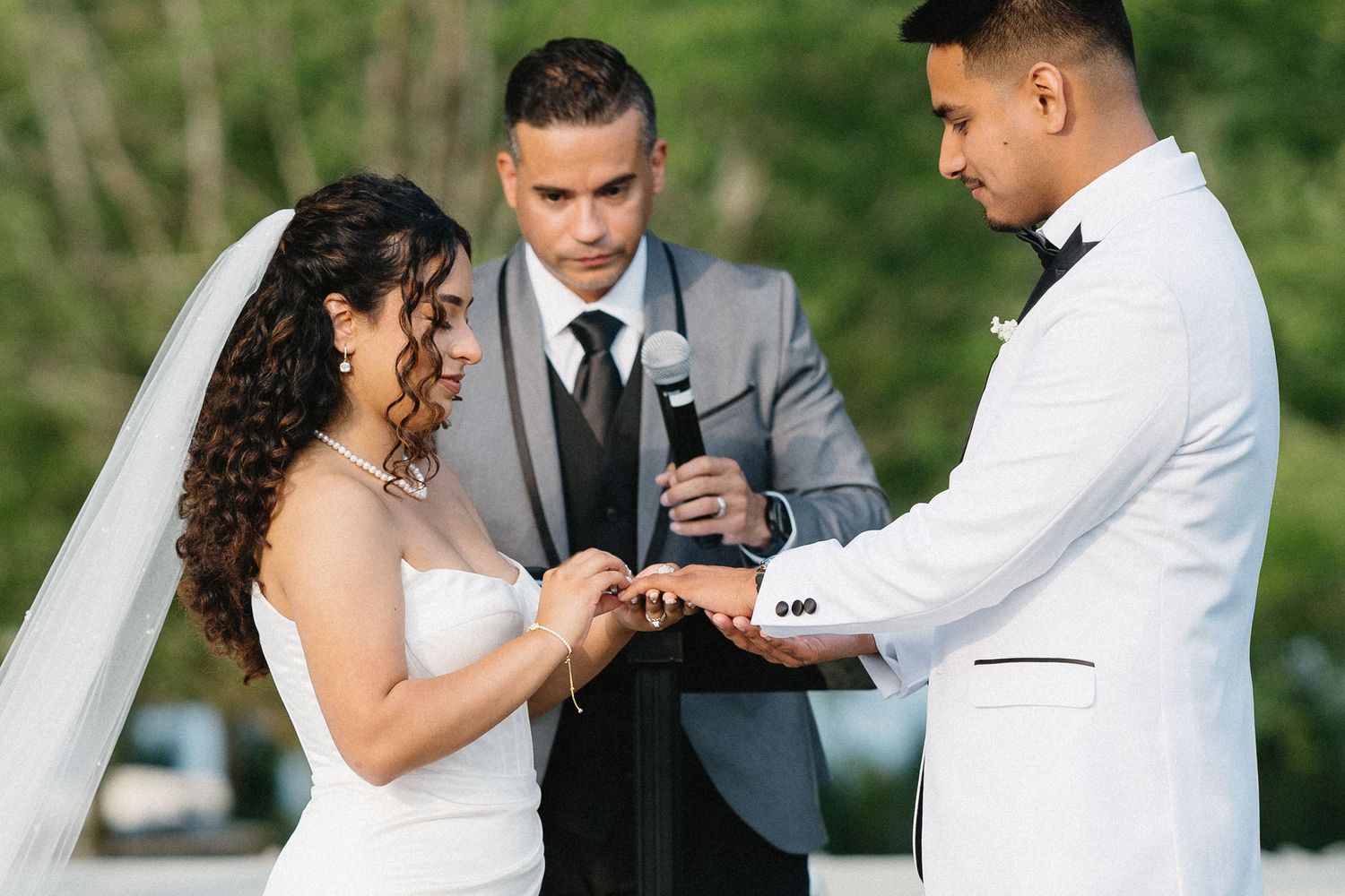 Bride and groom exchange rings during outdoor wedding ceremony with officiant.