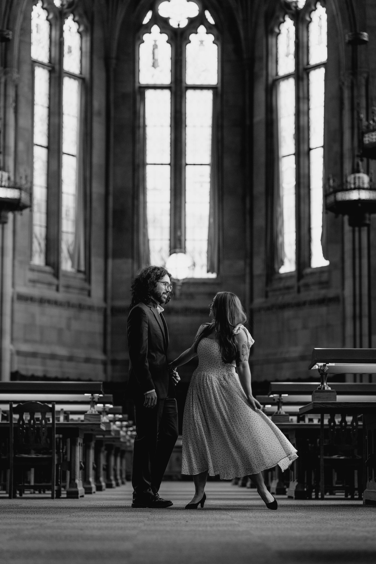 Black and white photo of a couple holding hands in a grand gothic cathedral with tall arched windows.