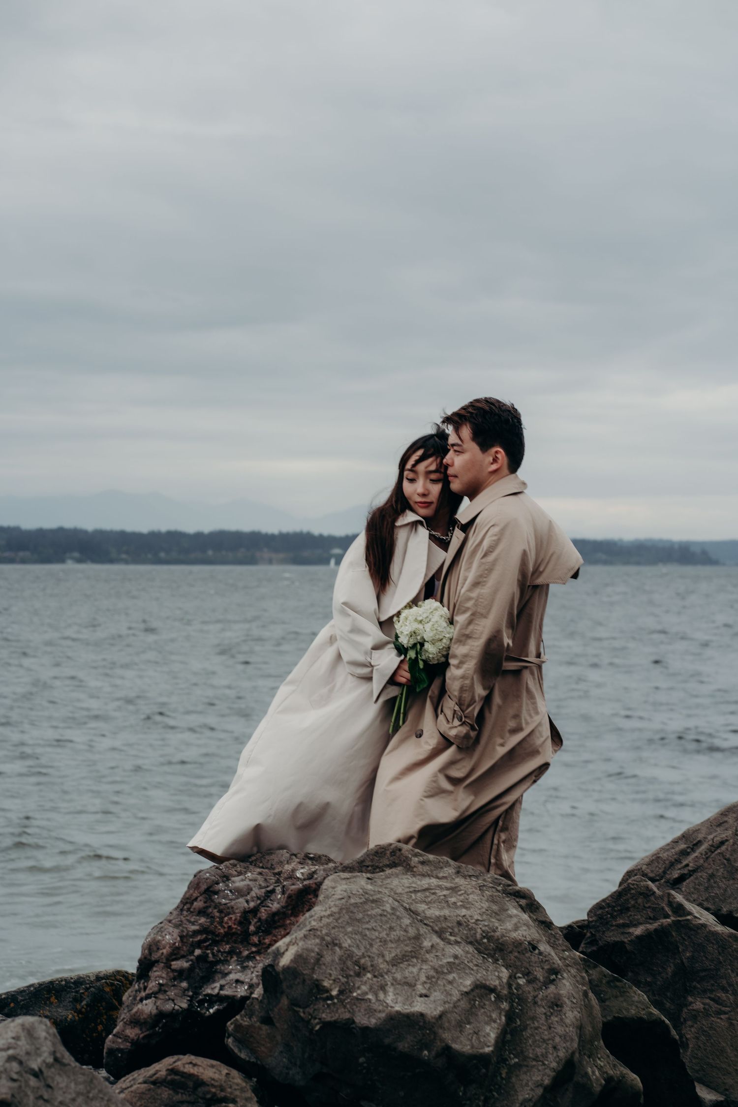 Couple in matching beige coats embracing on rocky shoreline with bouquet under overcast sky.