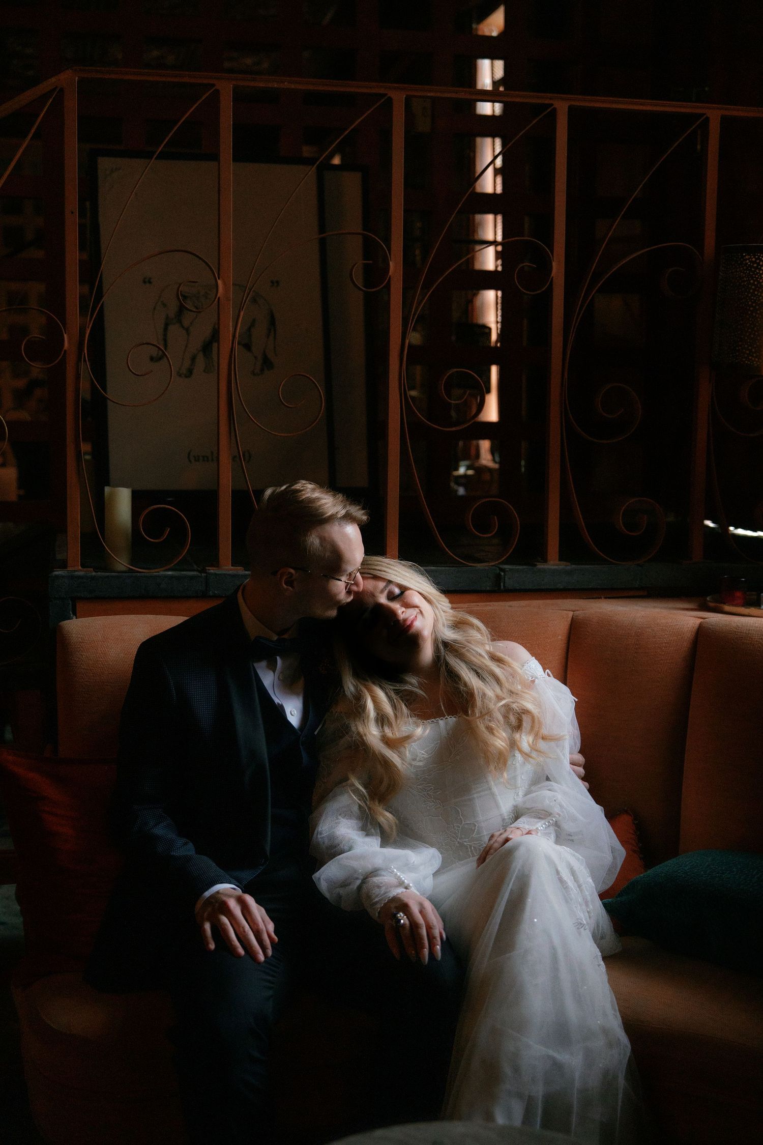 Bride in white gown rests head on groom in black tuxedo while seated on an orange velvet sofa.