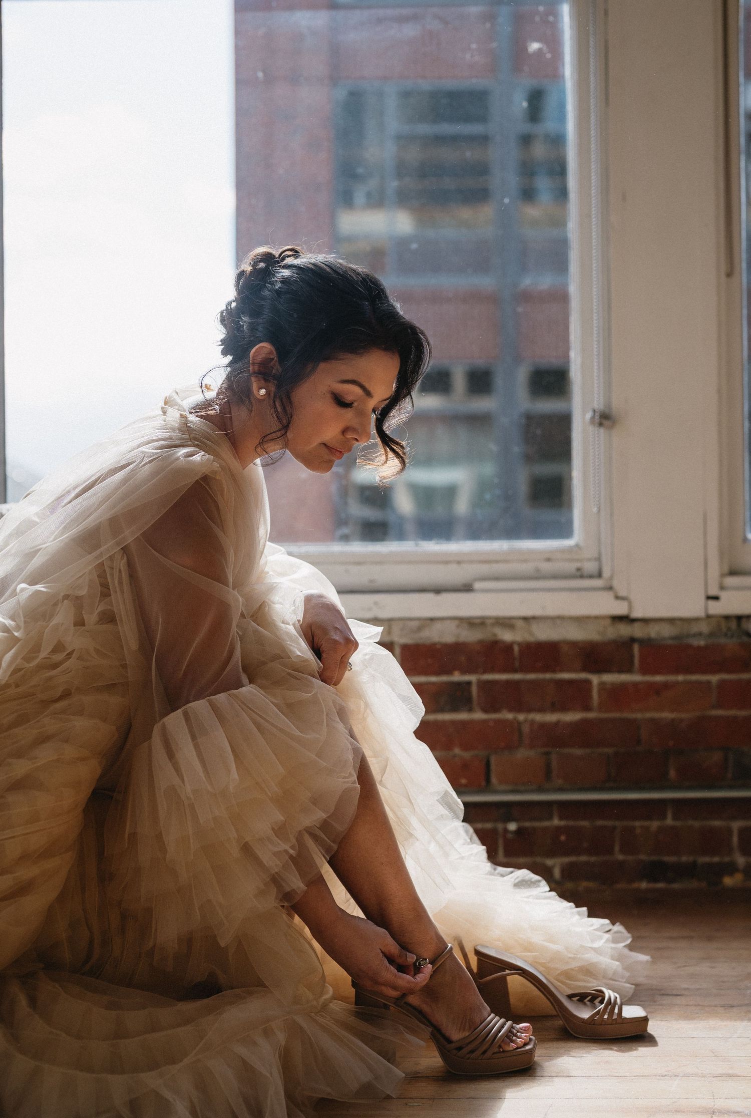 Bride in tulle gown fastens gold heels while seated by window with brick wall backdrop.