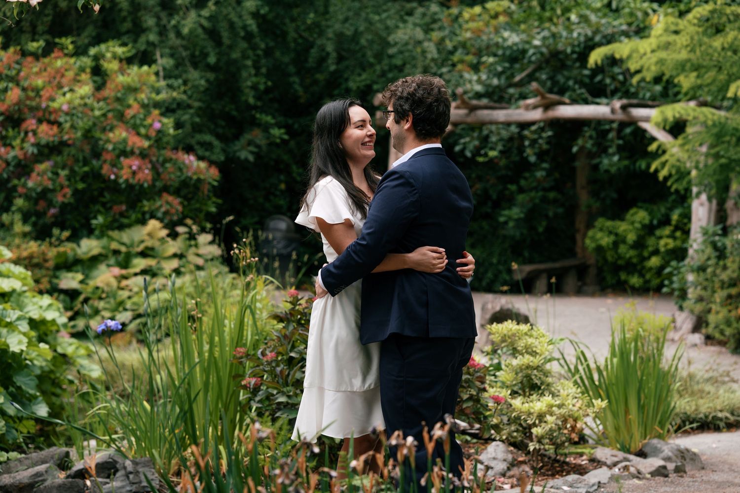 Happy couple embracing and smiling in a lush botanical garden setting, woman in white dress and man in navy suit.