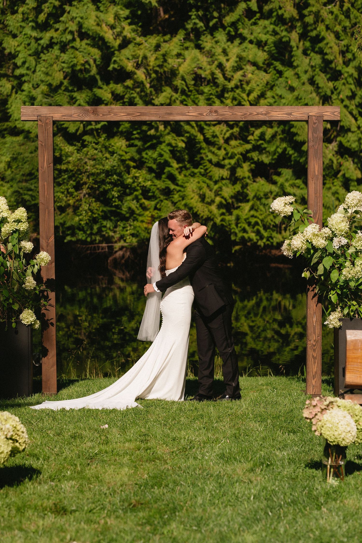 Bride and groom sharing first kiss under rustic wooden arch decorated with white hydrangeas.