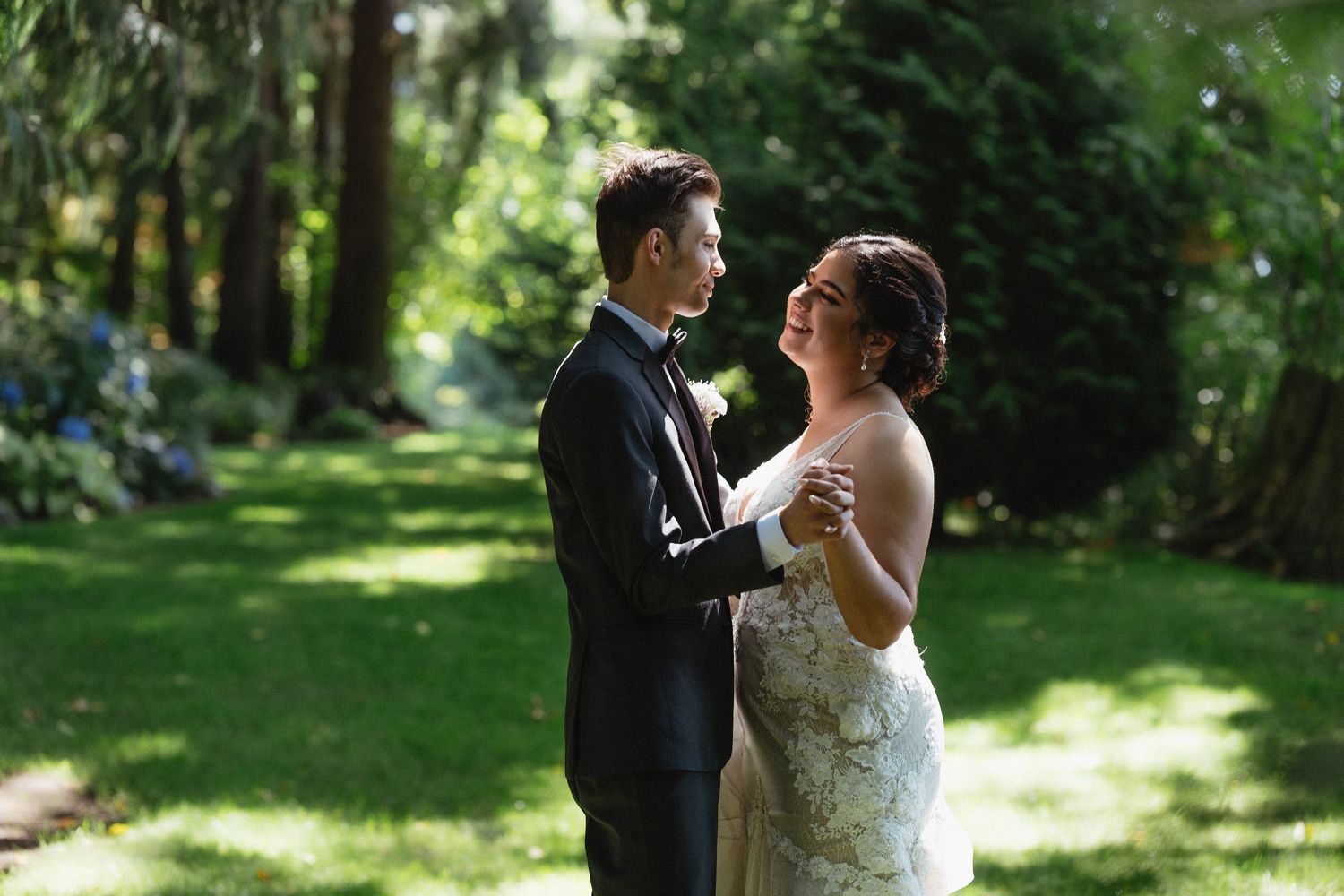 Bride and groom sharing a tender moment dancing together on lush green lawn in wooded setting.