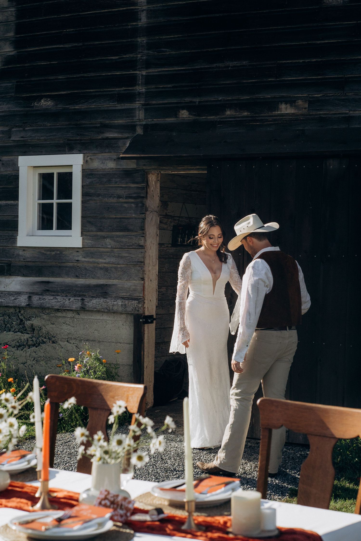 Bride in white gown and groom in cowboy hat hold hands outside a rustic dark wood barn.