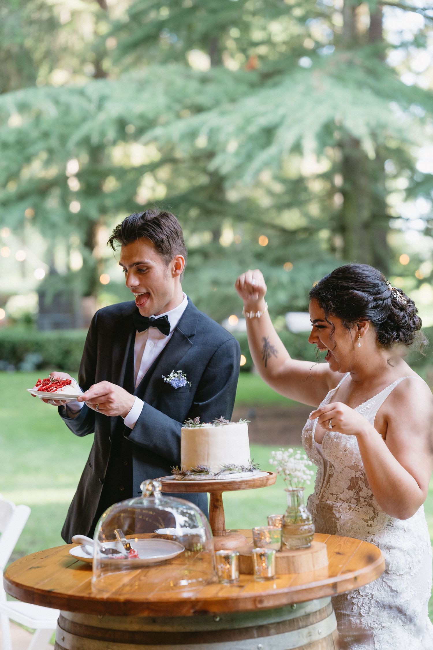 Excited bride and groom celebrating wedding cake cutting at rustic barrel dessert table outdoors.