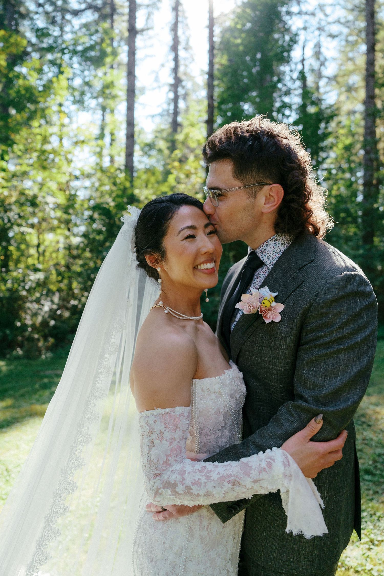 Groom kisses smiling bride on cheek as they embrace in sunlit forest setting.