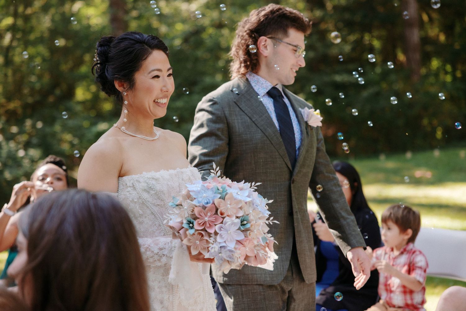 Bride and groom walk down the aisle surrounded by bubbles at an outdoor wedding ceremony.