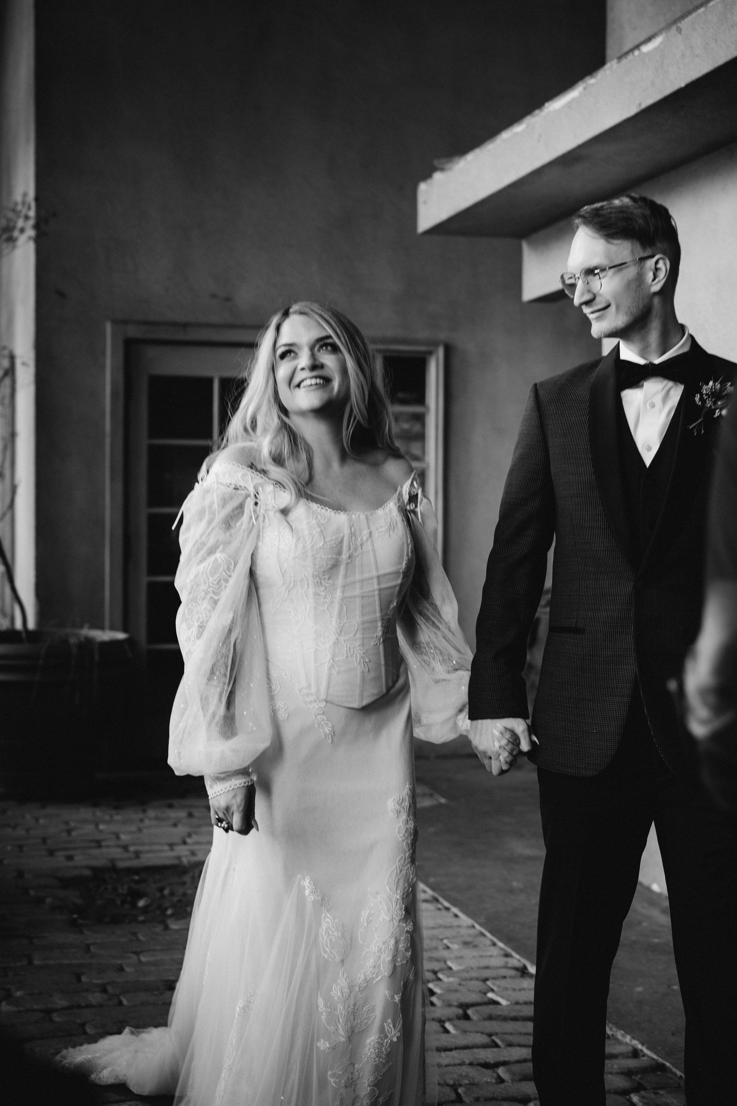 Black and white photo of smiling bride in lace gown with groom in tuxedo holding hands outdoors at wedding venue.