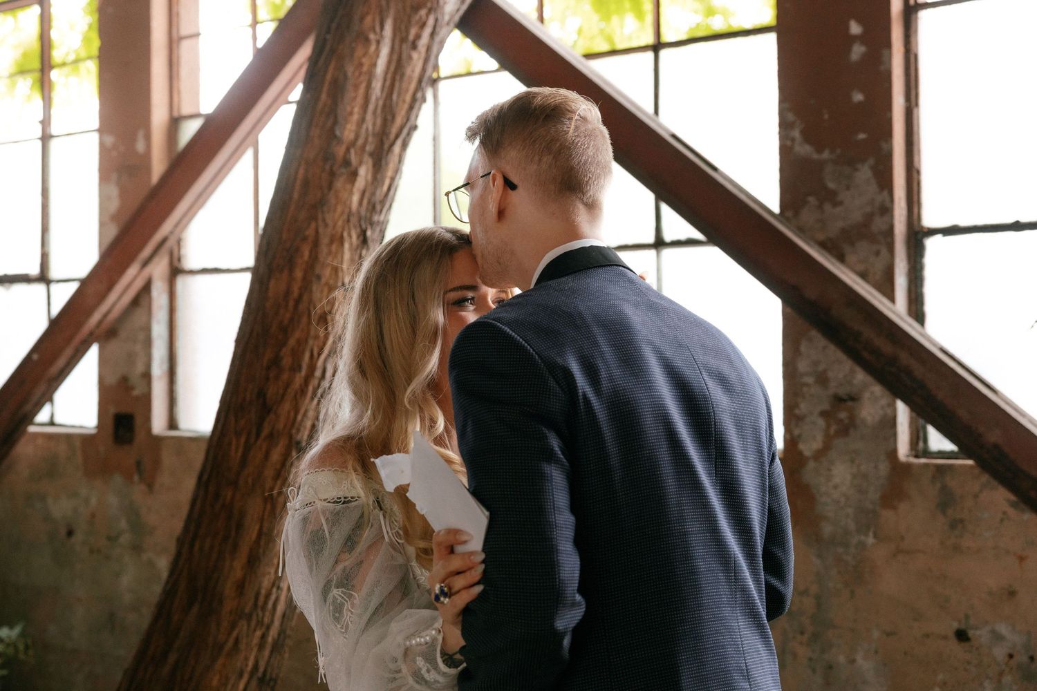Newlyweds share a tender kiss surrounded by exposed brick and wooden beams in an industrial wedding venue.