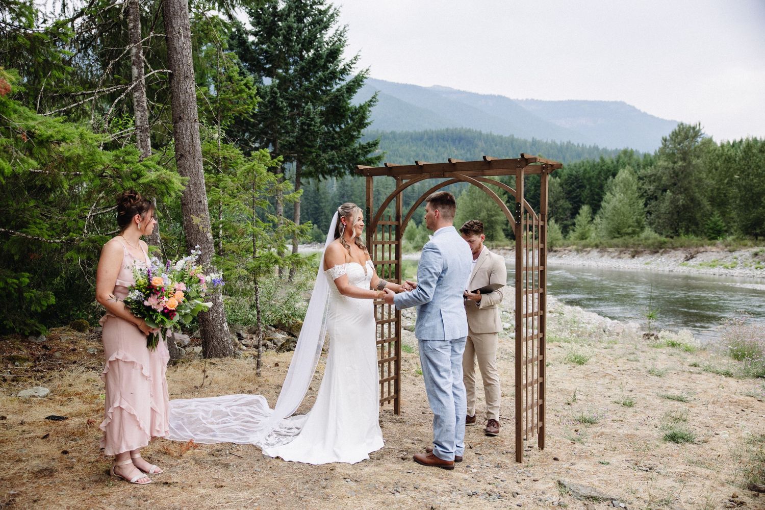 Outdoor wedding ceremony beside a river with couple exchanging vows under a wooden arch surrounded by trees and mountains.