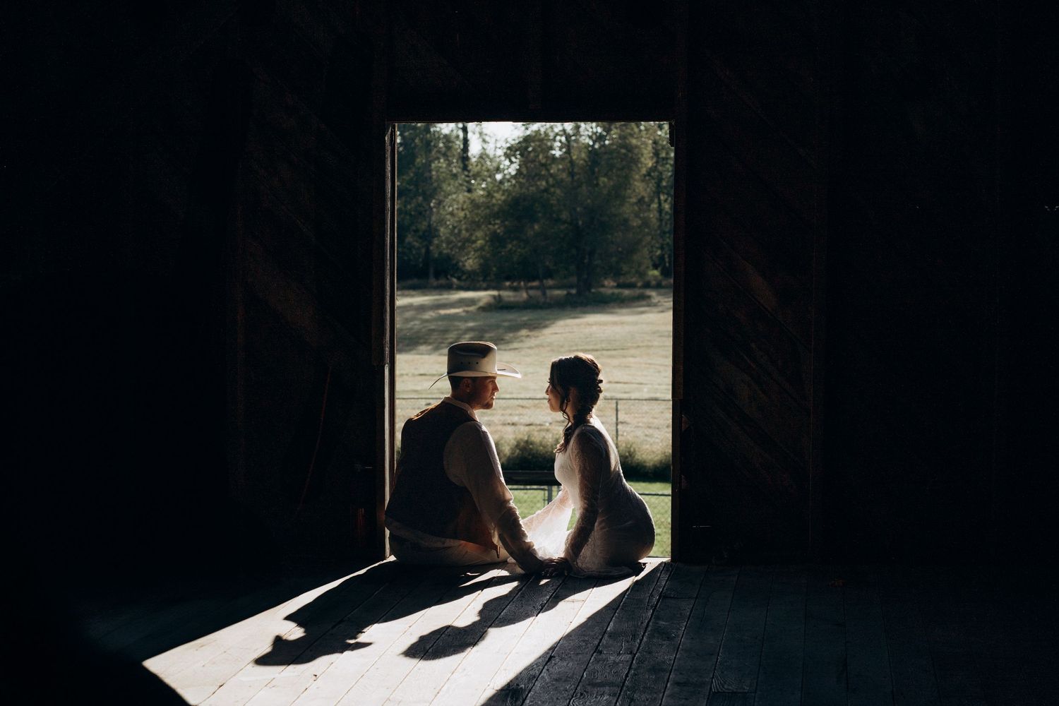 Silhouette of a couple sitting in a barn doorway overlooking a sunny green field.