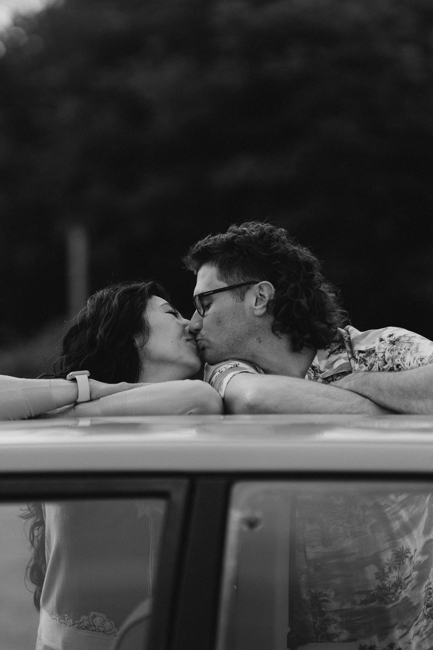 Black and white photo of a couple sharing a kiss over the roof of a vintage car at dusk.