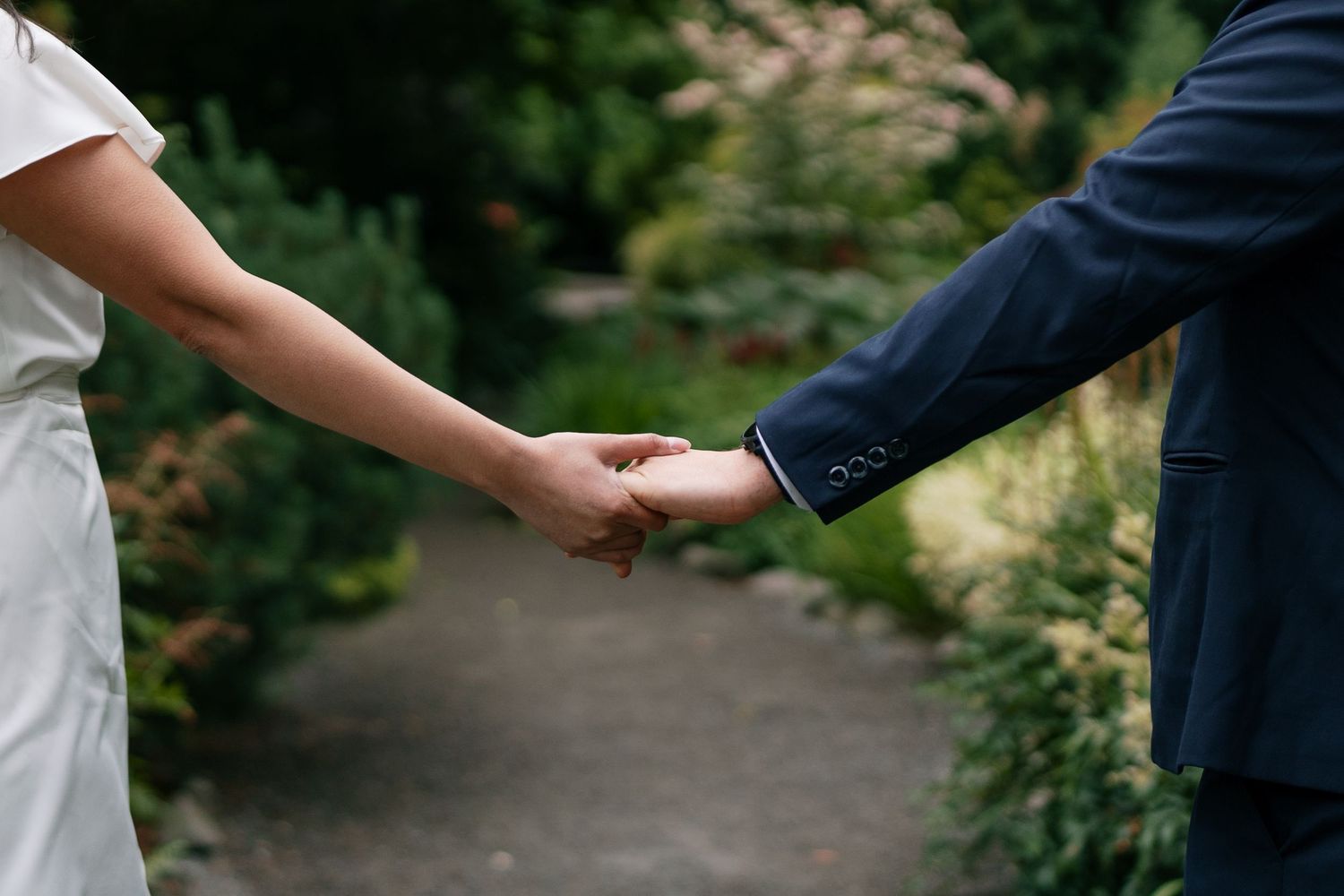Close-up of bride and groom holding hands while walking along a lush garden pathway.