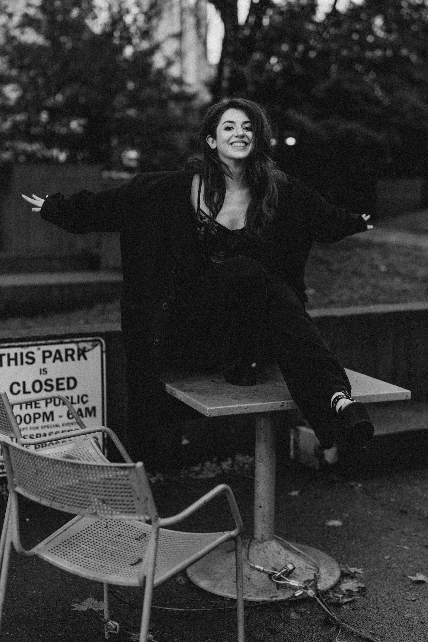 A joyful woman in a black coat sits on a park table with arms outstretched, smiling in a black and white photo.