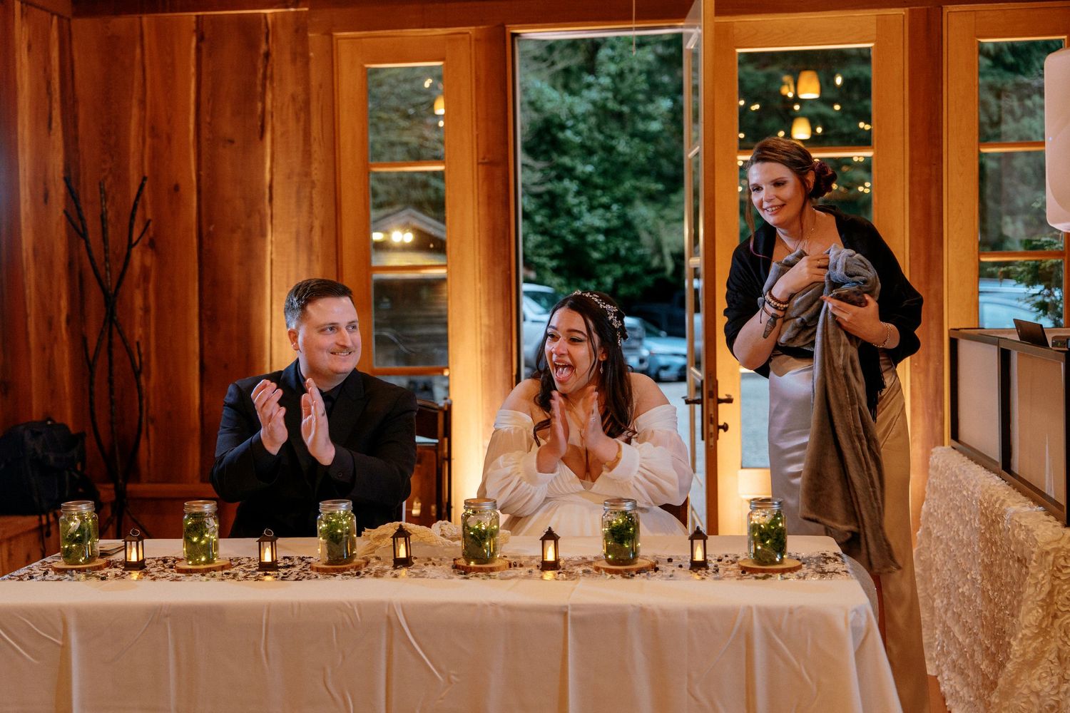 Bride and groom applaud at elegantly decorated head table with candles and greenery centerpieces.