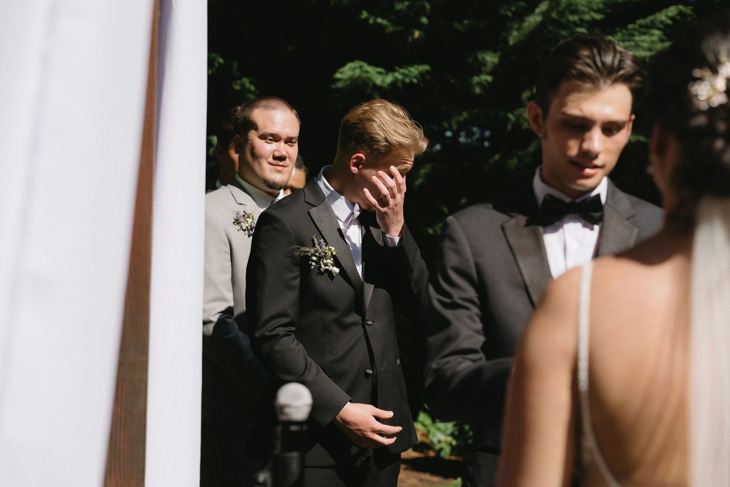 Emotional groomsman wipes tears during outdoor wedding ceremony while standing at the altar with wedding party.