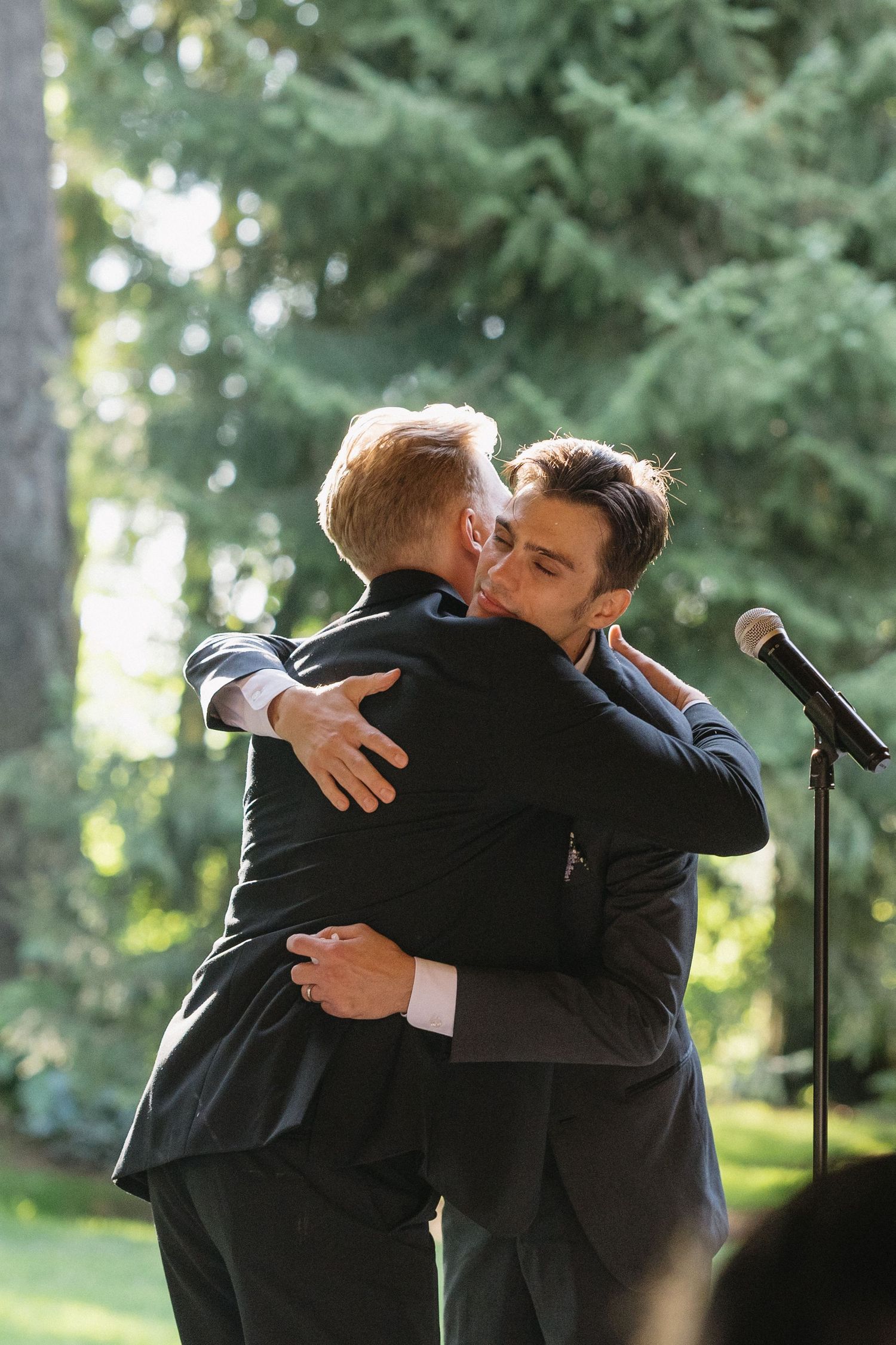 Two grooms in dark suits sharing an emotional hug near microphone at outdoor wedding ceremony.