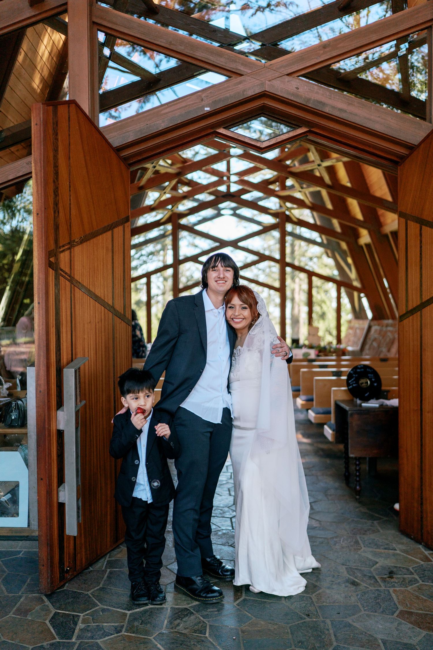 Bride and groom with young son posing at entrance of wooden A-frame wedding chapel.