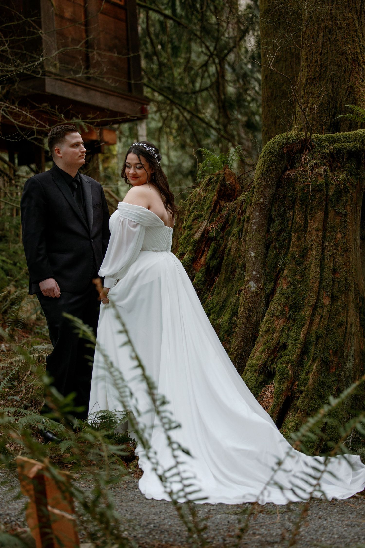 Bride in flowing white off-shoulder gown with groom beside a massive moss-covered tree in forest.
