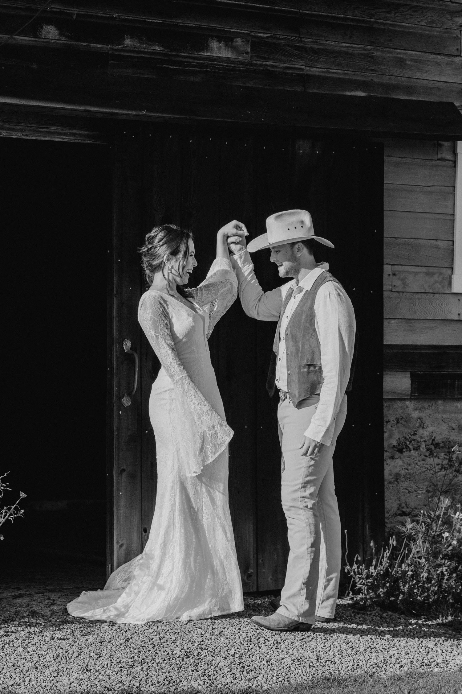 Black and white photo of bride and groom dancing outside rustic barn, groom in cowboy hat.