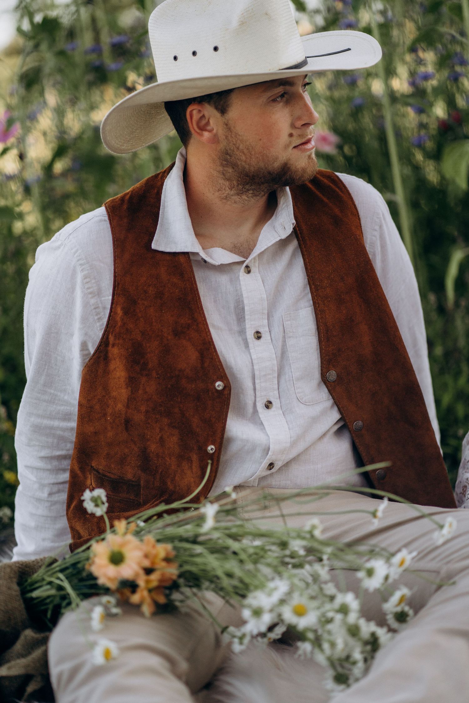 Handsome groom in white cowboy hat and brown suede vest sits outdoors surrounded by wildflowers.