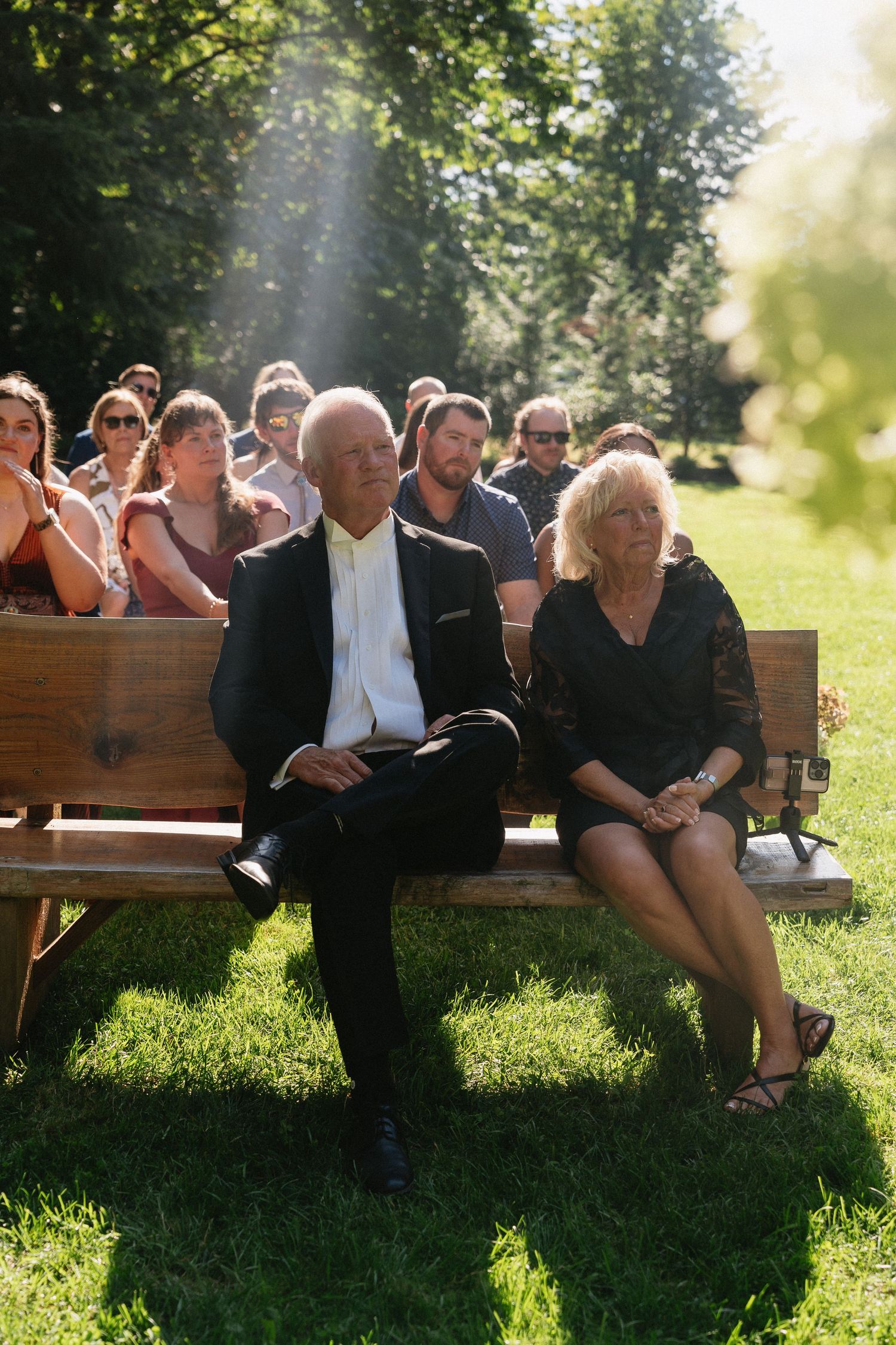 Wedding guests seated on wooden benches at outdoor ceremony in lush green garden setting.
