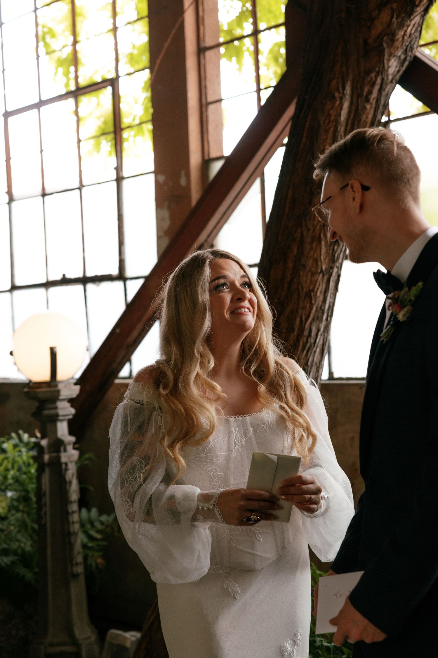 Radiant bride smiles while reading vows to groom in a sunlit industrial wedding venue.
