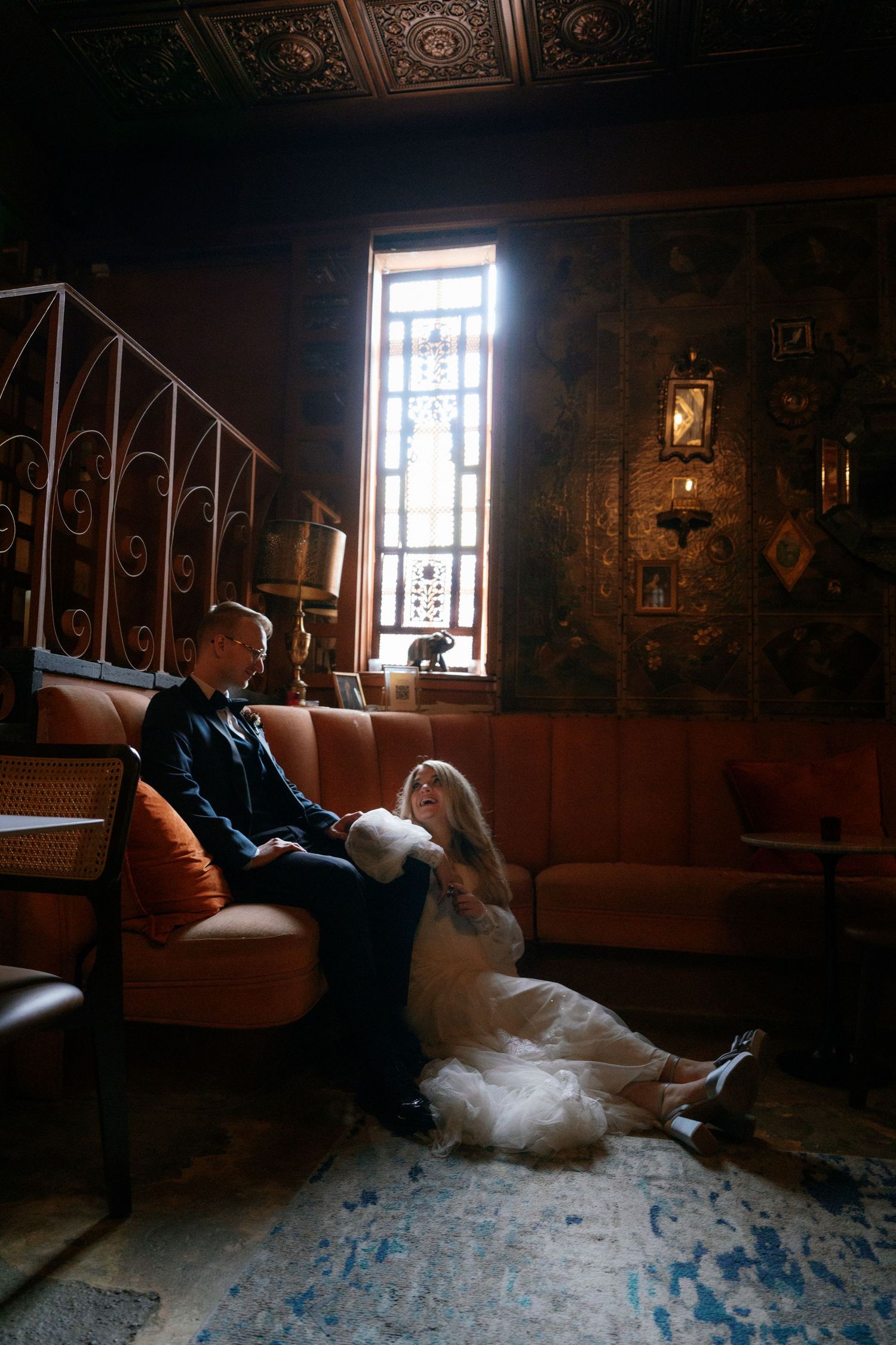 Bride in white gown sits on floor leaning against groom in dark suit in ornate vintage room with stained glass window.