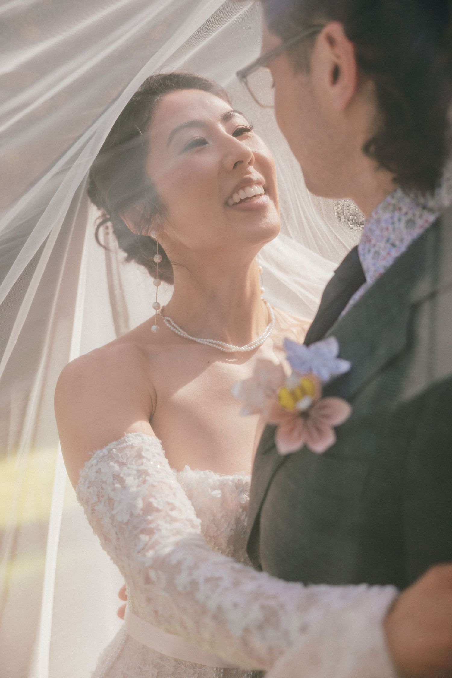 Smiling bride and groom share intimate moment beneath flowing wedding veil in sunlight.