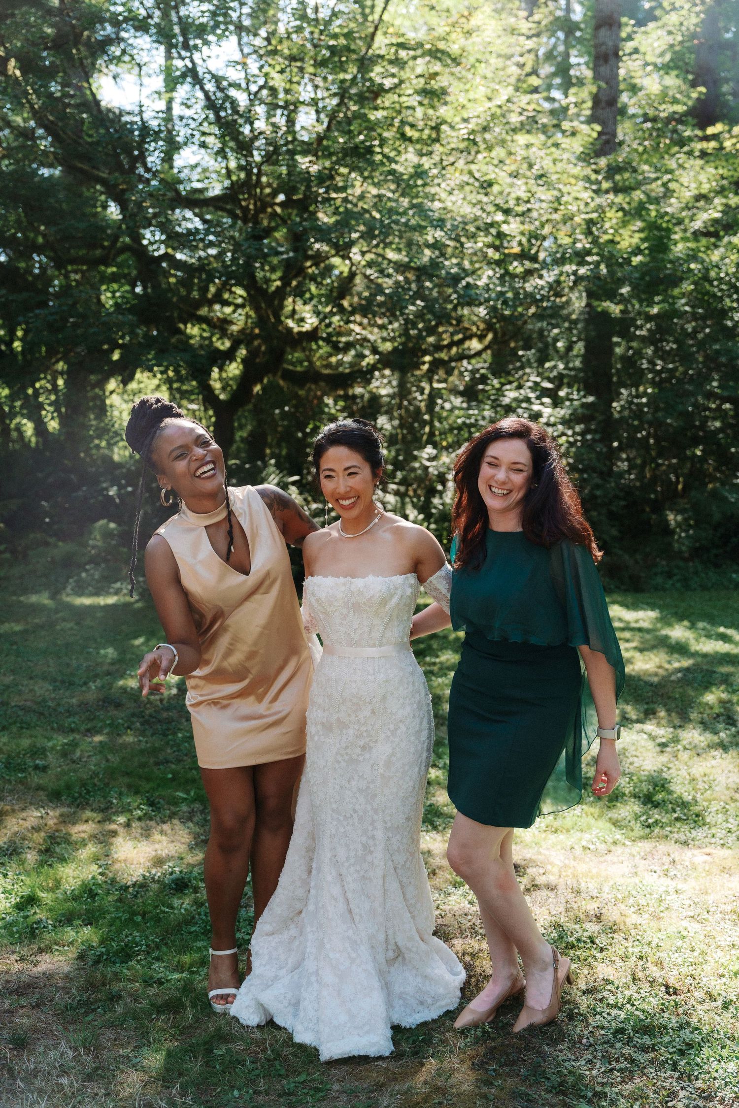 Bride in white lace gown laughs with two friends in gold and green dresses outdoors.