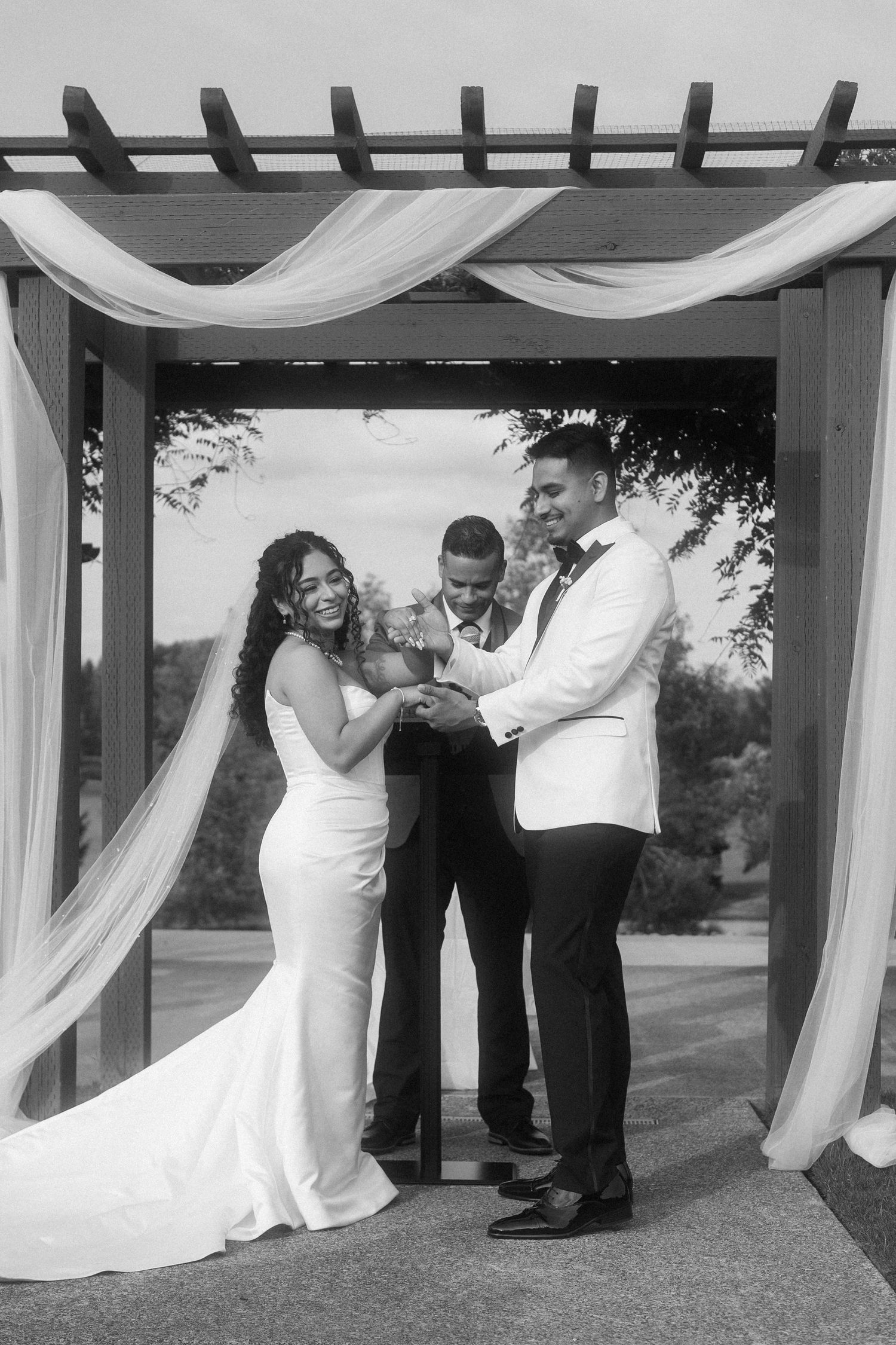 Black and white photo of a laughing bride and groom holding hands under a draped pergola at their wedding.
