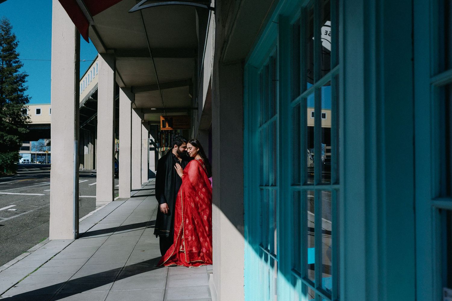 Couple in red and black traditional Indian outfits sharing a kiss in a sunlit urban colonnade.