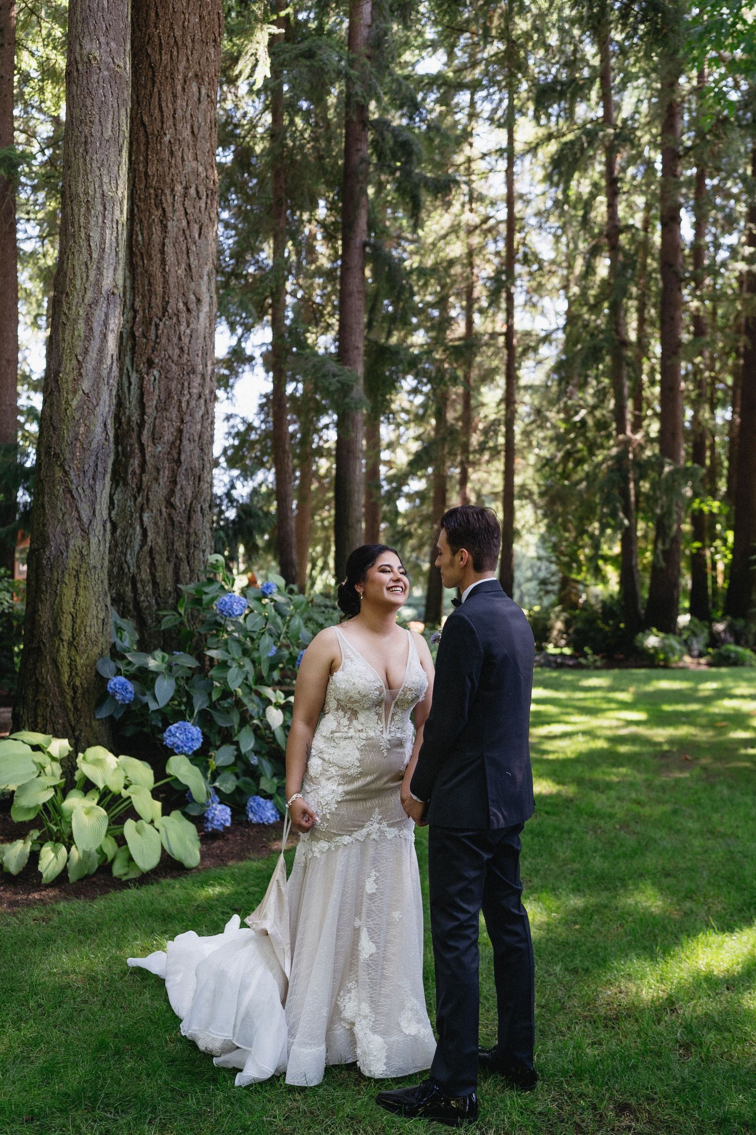 Bride in lace gown laughing joyfully with groom in black tux among tall trees and hydrangeas.