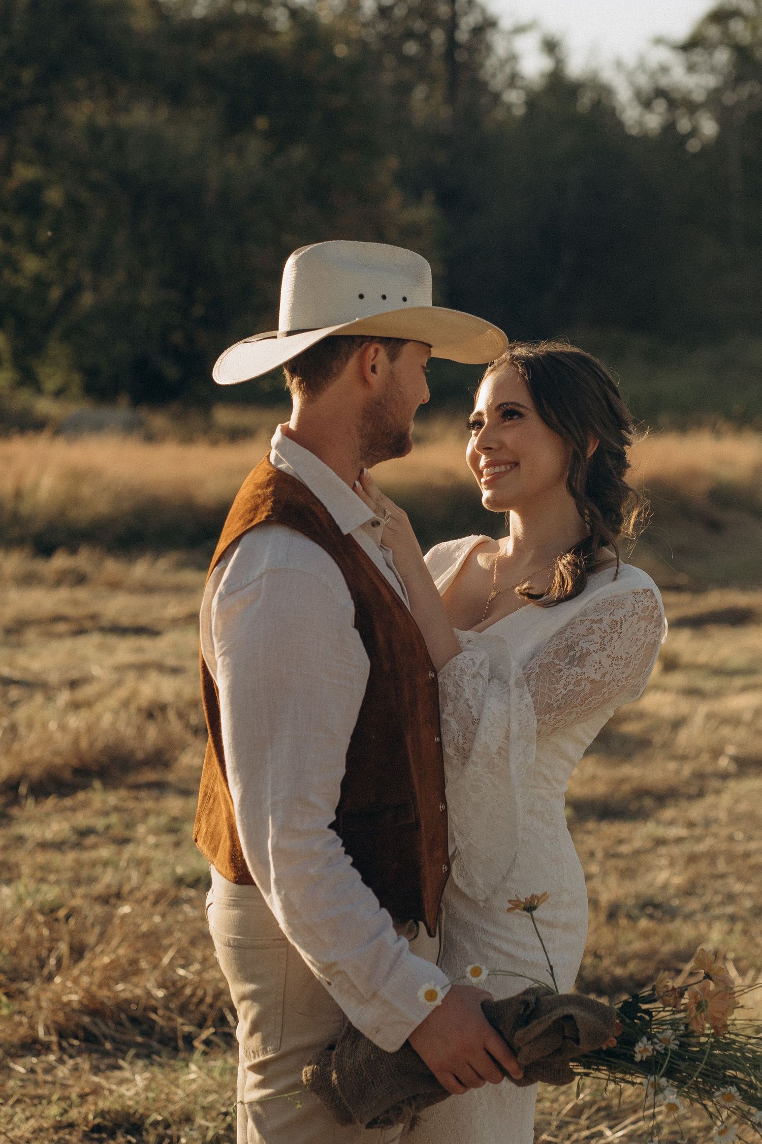 Bride smiling at cowboy hat groom in romantic rustic outdoor wedding portrait at sunset.