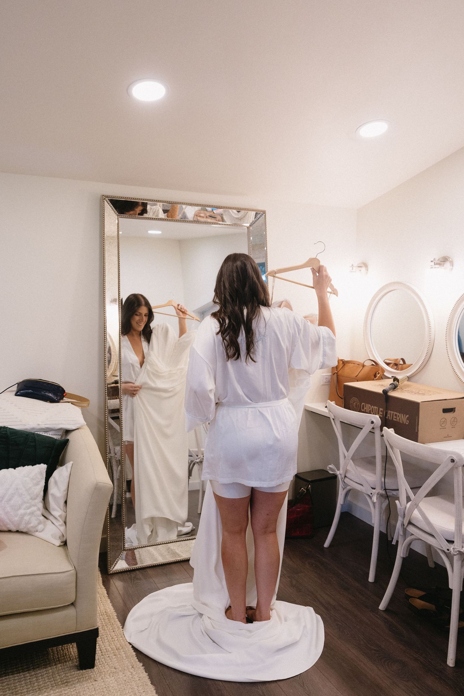 Bride in white robe holds her wedding dress on a hanger while viewing her reflection in a large floor mirror.
