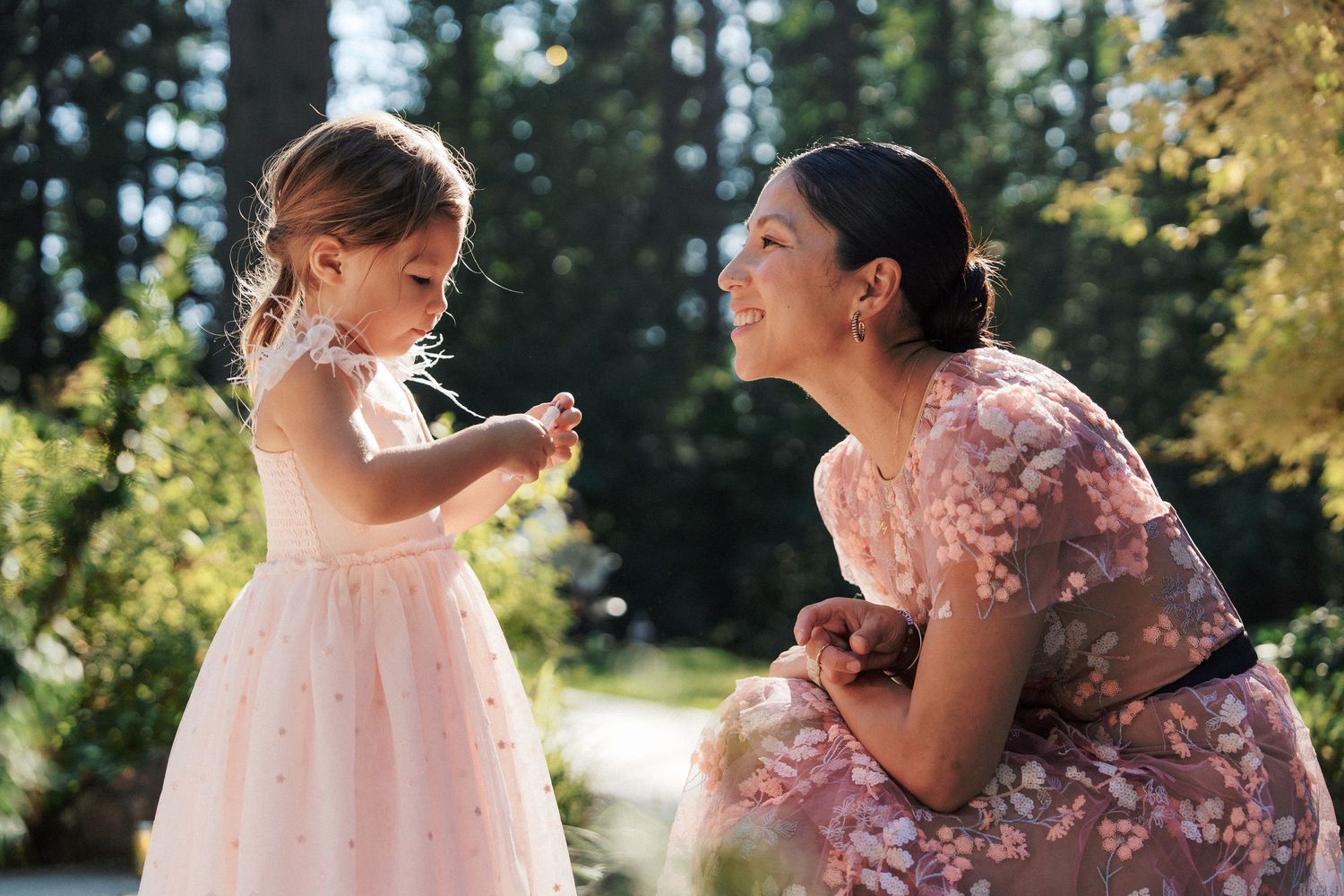 Woman in floral dress crouches to interact with little girl in pink tulle dress in garden.