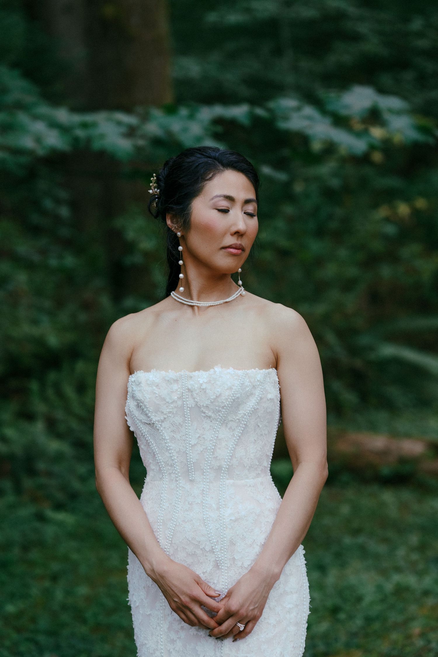 Serene bride in strapless white lace wedding gown with eyes closed poses in a lush green forest.