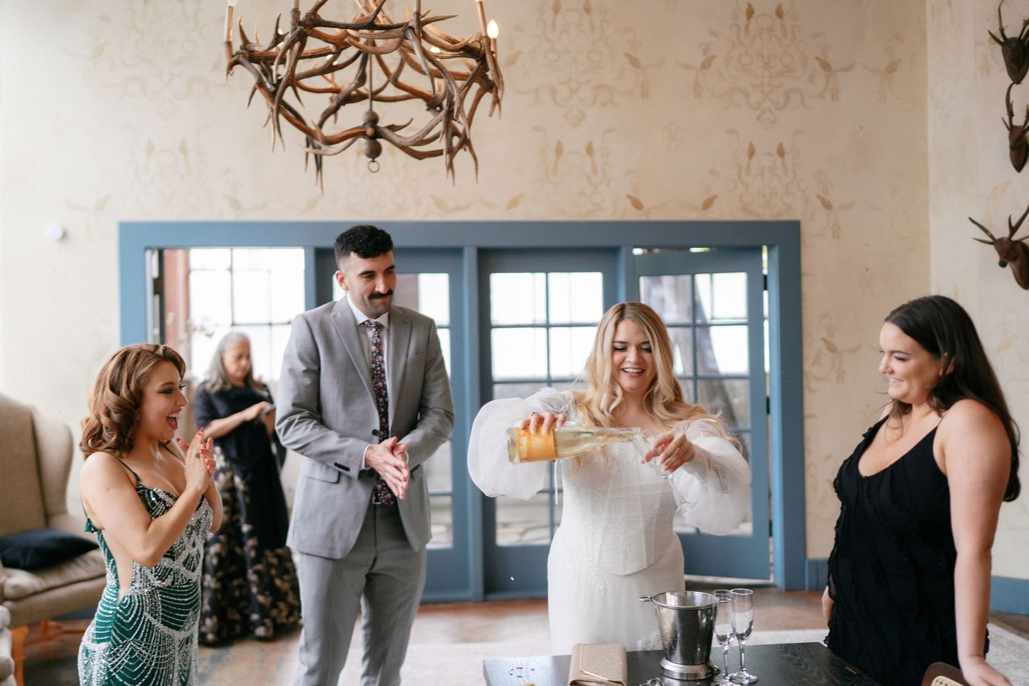 Bride in white dress pouring drinks with guests under antler chandelier at wedding venue.
