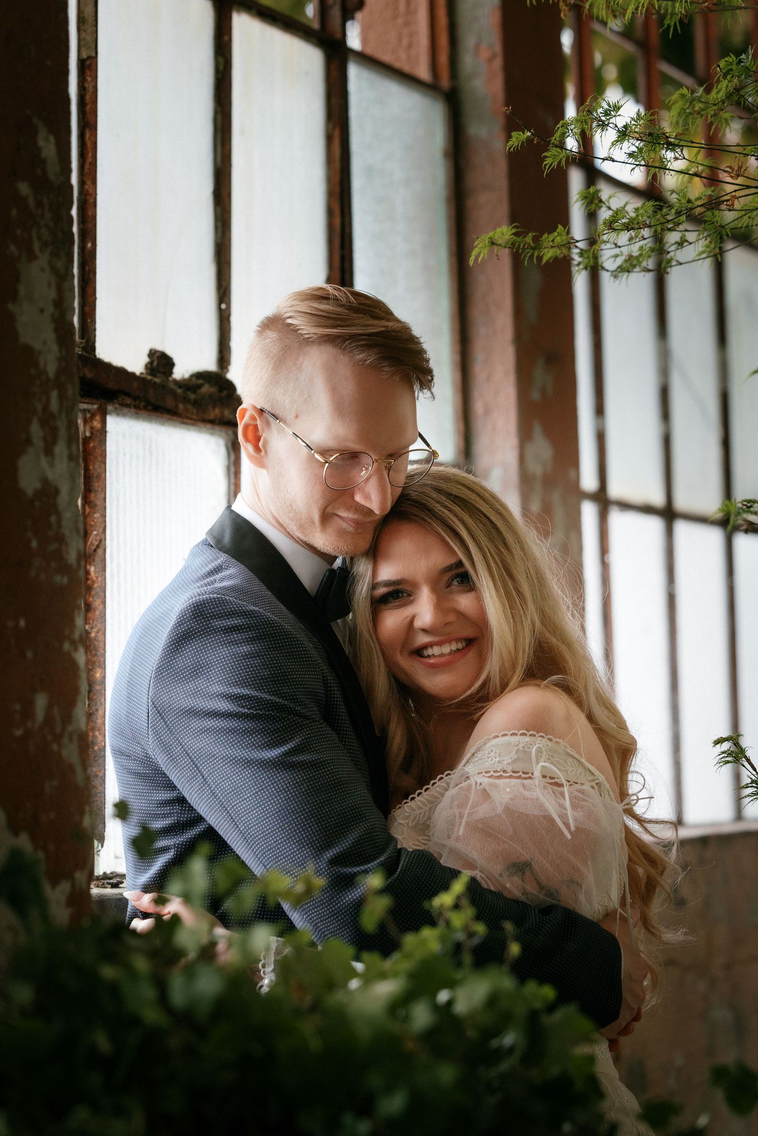 Smiling bride in off-shoulder lace gown embracing groom in glasses near a rustic industrial window.
