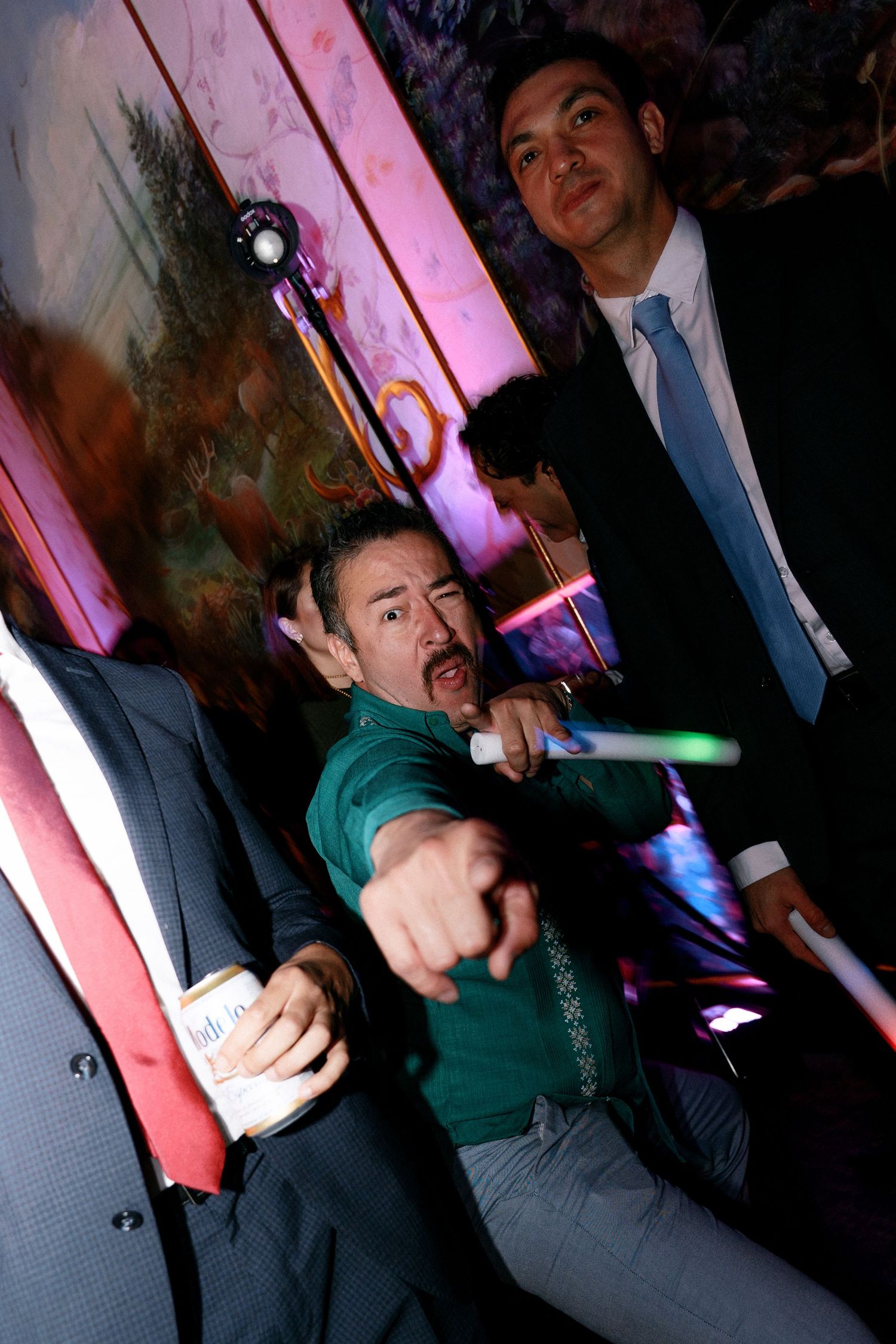 Man in green shirt pointing at camera with glow stick at wedding party.