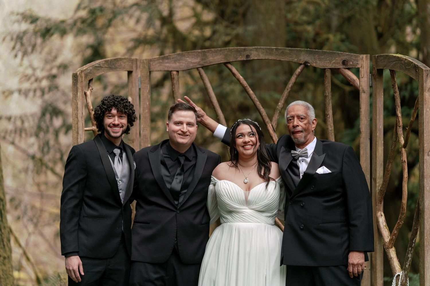 Wedding party group photo with bride in white gown and three men in black suits under wooden arch.
