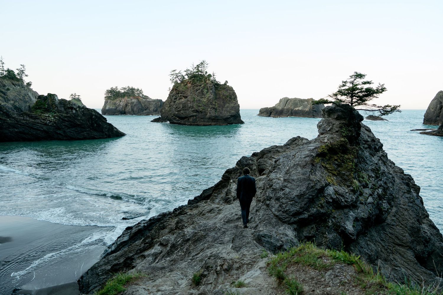 Person standing on rocky coastal cliffs with dramatic sea stacks and calm blue ocean water.