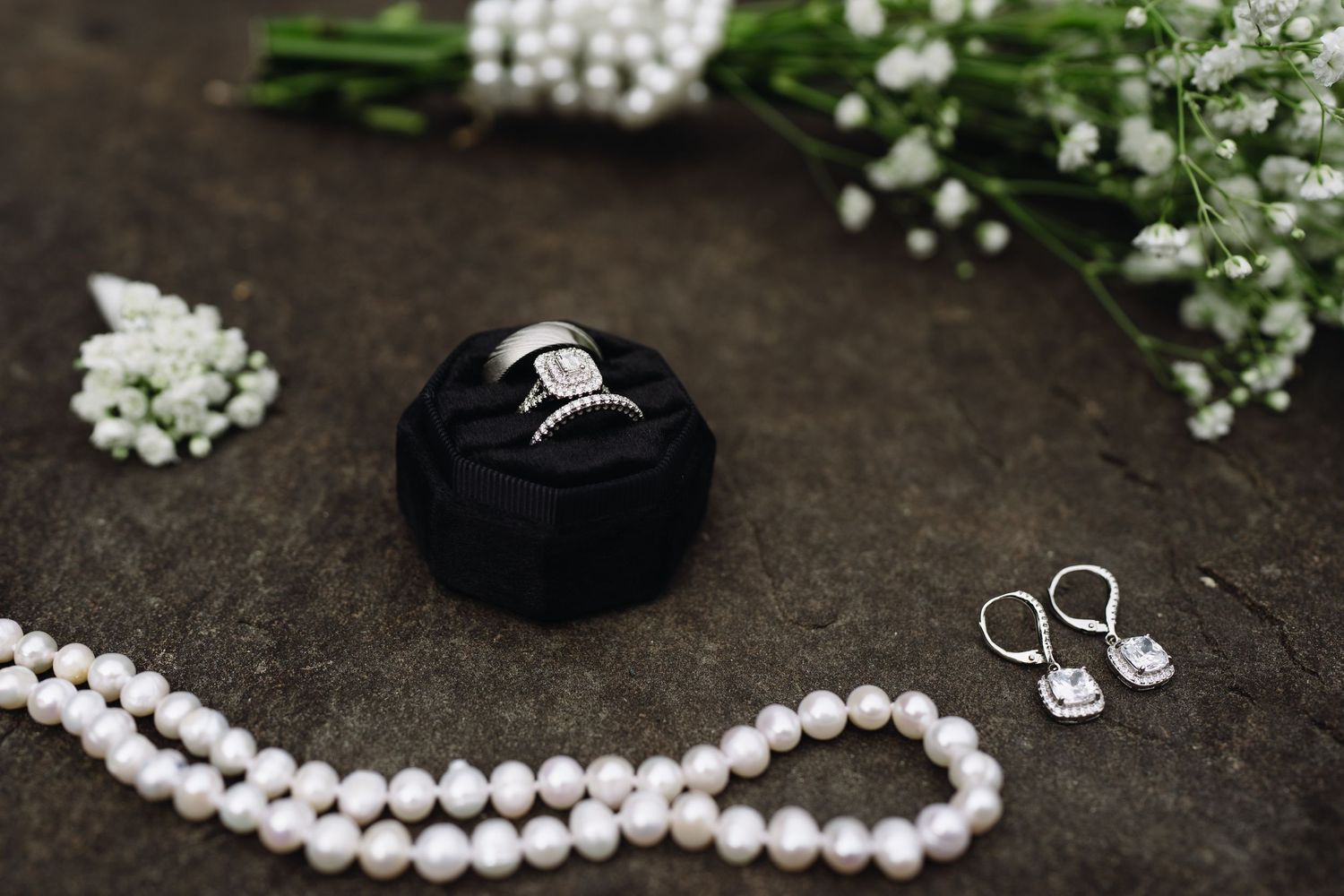 Wedding rings in black velvet box with pearl necklace, diamond earrings, and white flowers on stone.