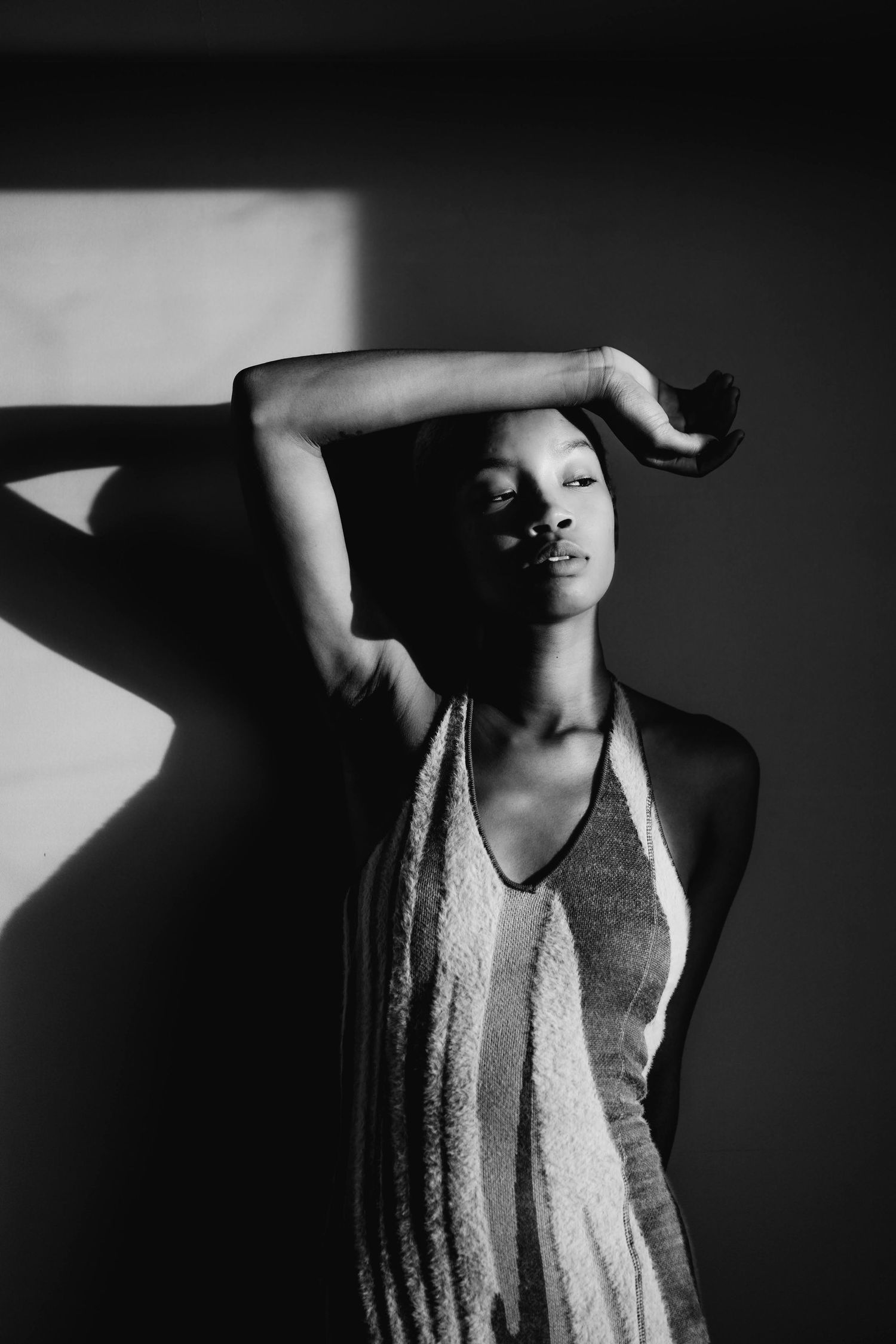 Black and white artistic portrait of a woman in a halter dress posing with arm raised against a shadowed wall.